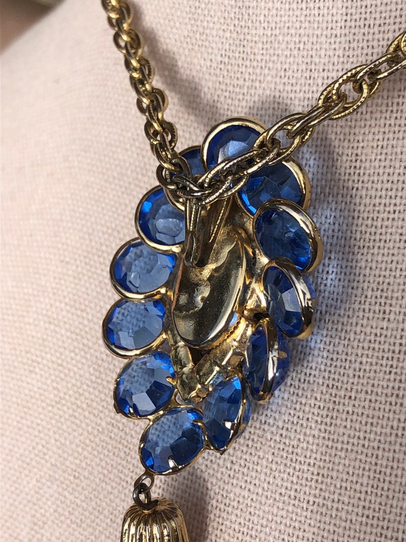May include: A gold chain necklace with a blue rhinestone pendant and a gold tassel. The pendant is shaped like a flower with multiple layers of rhinestones.