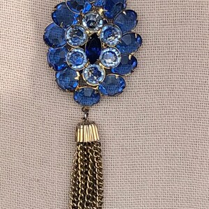May include: A gold chain necklace with a blue rhinestone pendant and a gold tassel. The pendant is a cluster of blue rhinestones in a flower shape.