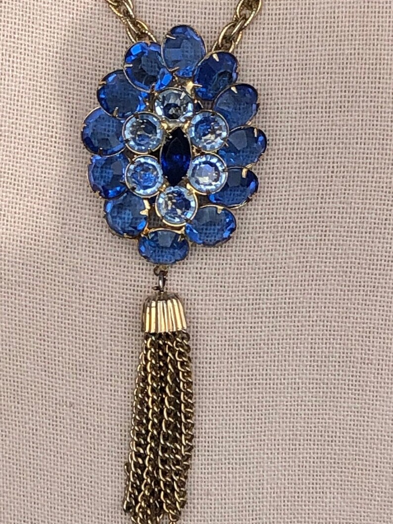 May include: A gold chain necklace with a blue rhinestone pendant and a gold tassel. The pendant is a cluster of blue rhinestones in a flower shape.
