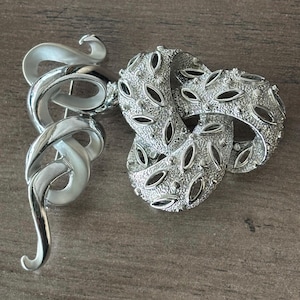 May include: A silver-toned brooch with a swirling, ribbon-like design and a textured, knotted section. The brooch has a polished finish and features oval cutouts in the knotted section. The pin is a vintage accessory.