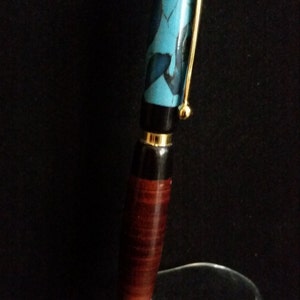 Puede incluir: Un bolígrafo artesanal con clip y detalles dorados. El bolígrafo presenta una parte superior con estampado turquesa y negro, un centro negro y un barril de madera marrón rojizo. El bolígrafo se exhibe sobre un soporte transparente sobre un fondo oscuro.