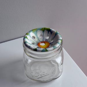 Puede incluir: Un tarro de vidrio transparente Ball con una tapa con estampado de margaritas. La tapa presenta una margarita blanca con un centro amarillo y hojas verdes. El tarro está sobre una superficie blanca.