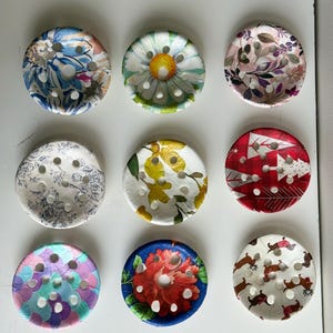 Pode incluir: Nove botões redondos decorativos com padrões florais. Os botões apresentam vários desenhos, incluindo margaridas, folhas e árvores de Natal. Cada botão tem vários orifícios para costurar e é feito de um material semelhante a papel.