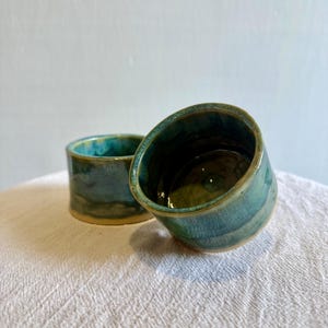 Pode incluir: Duas canecas de cerâmica feitas à mão com esmalte turquesa e azul-petróleo. As canecas têm formato cilíndrico com uma borda ligeiramente angulada. O esmalte tem um acabamento brilhante e uma aparência texturizada. As canecas são exibidas em uma superfície branca texturizada.