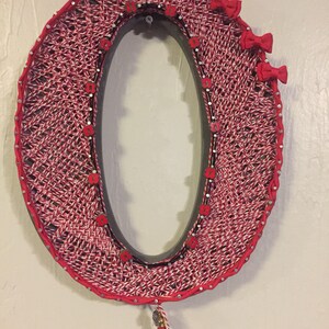 May include: Oval-shaped wall decor featuring a woven red and white pattern. The frame is outlined with red trim and adorned with small red bows, buttons, and a tassel. Silver studs secure the woven design.