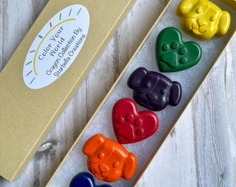 May include: A box of colorful crayons in the shape of dogs and hearts. The crayons are yellow, green, purple, red, orange, and blue. The box has the text "Color Your World Crayon Collection By Starbella Creations."