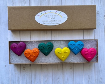 May include: A set of six heart-shaped crayons in a cardboard box. The crayons are purple, orange, green, blue, and pink. Each crayon has a different face on it. The box has a label that says "Color Your World Crayon Collection By Starbella Creations."