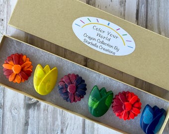 May include: A box of flower-shaped crayons in various colors, including orange, yellow, dark red, green, red, and blue. The crayons are arranged inside a tan cardboard box. The box has a white oval sticker with the text "Color Your World Crayon Collection By Starbella Creations."