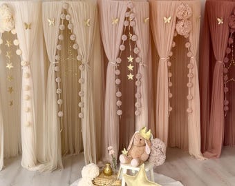 May include: Five sheer fabric bed canopies in shades of cream, beige, light pink, dusty rose, and terracotta. Each canopy features hanging pom-pom garlands, gold butterfly accents, and star-shaped fairy lights. A plush rabbit toy sits on a small stool.