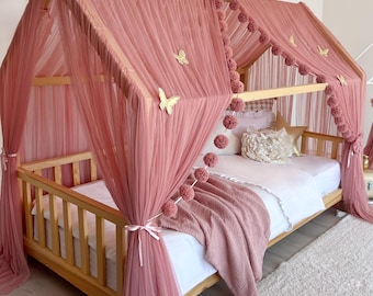 Op de afbeelding: Een houten bedframe in de vorm van een huis, gedrapeerd met oudroze tule. De hemel is voorzien van gouden vlinderaccenten en een slinger van bijpassende pompons. Het bed is opgemaakt met wit beddengoed, een oudroze textuurdeken en sierkussens.