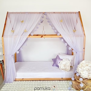 Könnte beinhalten: Ein hölzerner, hausförmiger Bettrahmen mit einem Baldachin aus lilafarbenem Tüll und Pomponbesatz. Das Bett hat ein weißes Laken und ein lilafarbenes Kissen mit einem goldenen Stern. Der Baldachin ist mit goldenen Schmetterlingen und Pompons verziert.