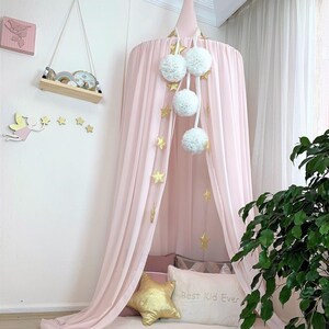 Puede incluir: Una cama con dosel rosa con pompones blancos y decoraciones de estrellas doradas. El dosel est&aacute; drapeado sobre una cama con almohadas, incluyendo una almohada dorada en forma de estrella y una almohada blanca con el texto "Best Kid Ever."