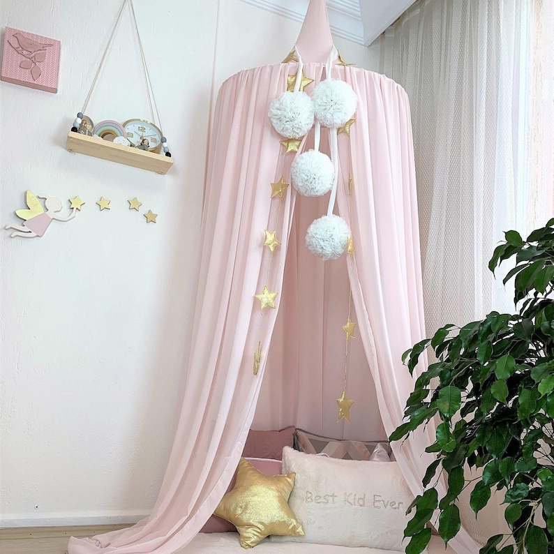 Puede incluir: Una cama con dosel rosa con pompones blancos y decoraciones de estrellas doradas. El dosel est&aacute; drapeado sobre una cama con almohadas, incluyendo una almohada dorada en forma de estrella y una almohada blanca con el texto "Best Kid Ever."