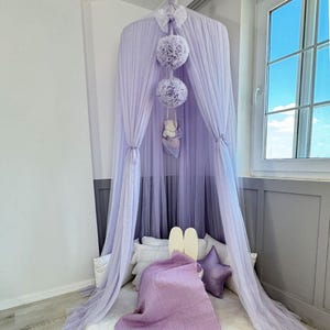 May include: A lavender tulle canopy with three fluffy pom-poms hanging from the top. The canopy is draped over a bed with white pillows and a purple blanket.