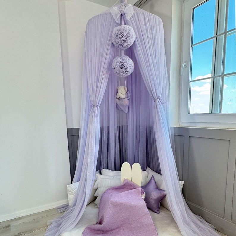May include: A lavender tulle canopy with three fluffy pom-poms hanging from the top. The canopy is draped over a bed with white pillows and a purple blanket.