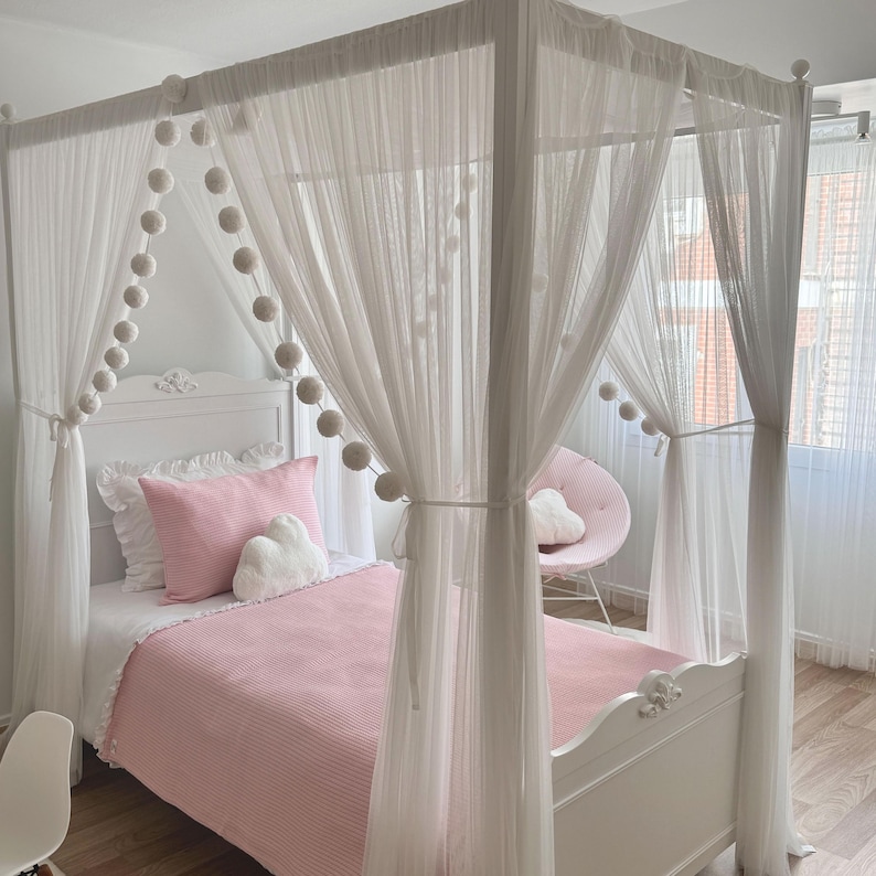 May include: A white canopy bed with a pink blanket and white pillows. The bed has a white canopy with white pom poms around the edges.