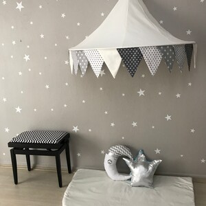 K&ouml;nnte beinhalten: Ein wei&szlig;es Stoff-Baldachin-Zelt mit einem grauen und wei&szlig;en Muster am Rand h&auml;ngt von der Decke. Eine wei&szlig;e Spielmatte mit einem grauen und wei&szlig;en Halbmondkissen und einem silbernen Sternkissen liegt auf dem Boden. Ein schwarz-wei&szlig; gemusterter Hocker befindet sich im Hintergrund.