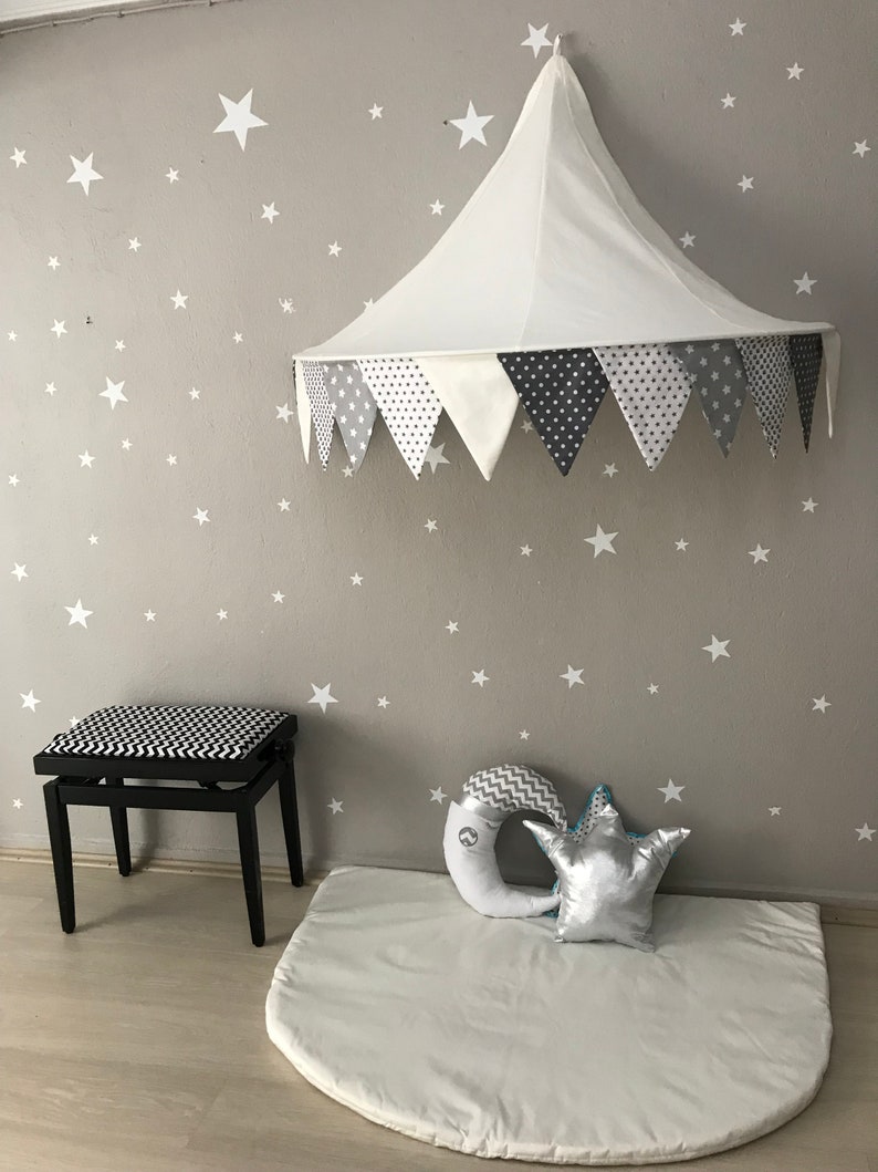 K&ouml;nnte beinhalten: Ein wei&szlig;es Stoff-Baldachin-Zelt mit einem grauen und wei&szlig;en Muster am Rand h&auml;ngt von der Decke. Eine wei&szlig;e Spielmatte mit einem grauen und wei&szlig;en Halbmondkissen und einem silbernen Sternkissen liegt auf dem Boden. Ein schwarz-wei&szlig; gemusterter Hocker befindet sich im Hintergrund.