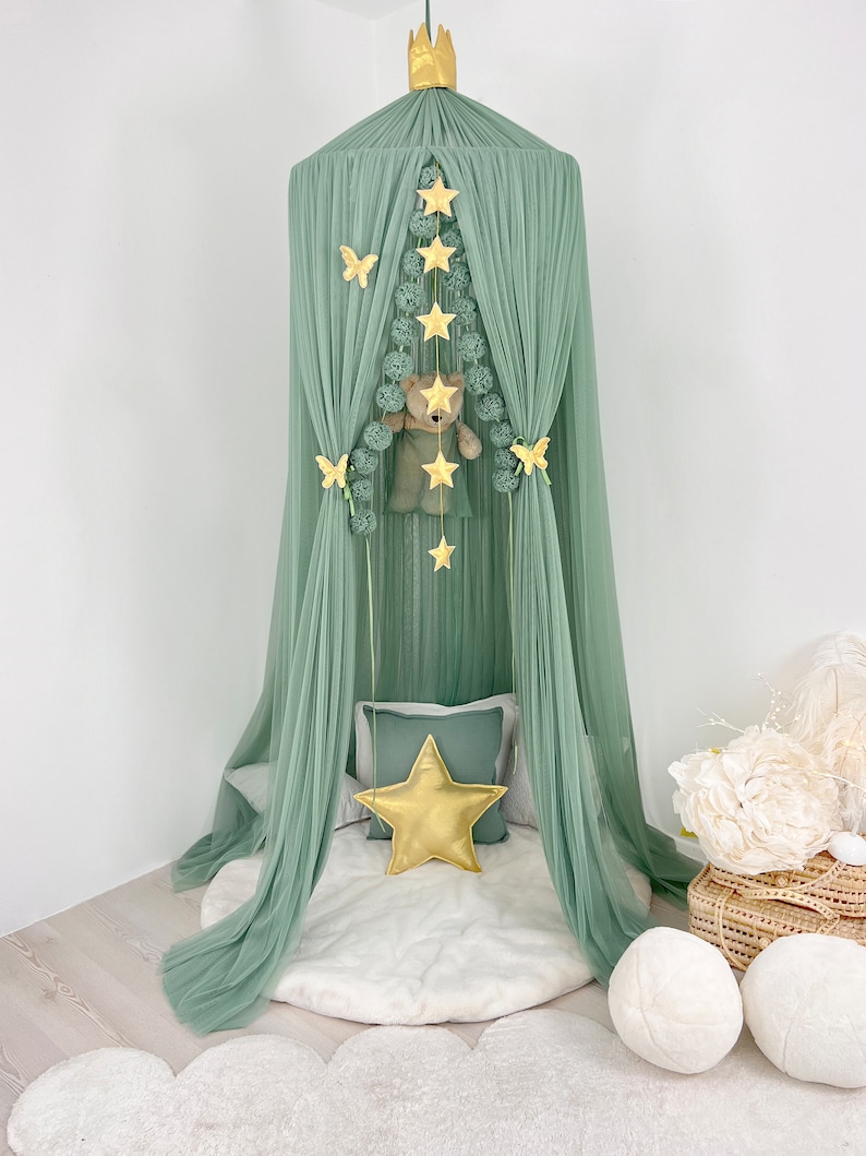 Puede incluir: Una carpa de tul verde con una corona dorada en la parte superior. La carpa est&aacute; decorada con estrellas doradas, mariposas y pompones. Un oso de peluche est&aacute; colgado dentro de la carpa.