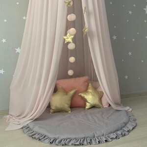 Puede incluir: Un dosel rosa p&aacute;lido enmarca una alfombra redonda gris con volantes. Cojines dorados en forma de estrella y guirnaldas de luces cuelgan encima. Una almohada rosa esponjosa y una pared gris con estrellas completan la escena.
