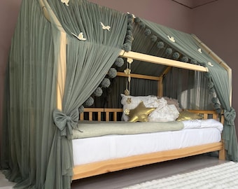 May include: A wooden house-shaped bed frame draped with sage green tulle fabric, featuring decorative white butterfly cutouts and a string of dark green pom-poms. The bed includes a white mattress, a gold star-shaped pillow, and white cushions.