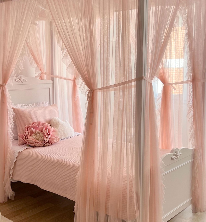 May include: A white canopy bed with a pink ruffled bedspread and pink sheer curtains. The bed has a white headboard with a carved design.