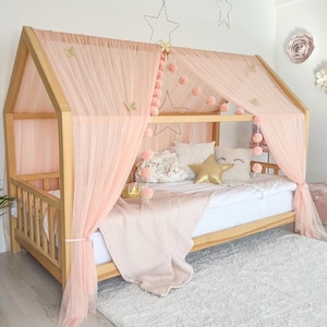 May include: A wooden house-shaped bed frame with a light pink tulle canopy. The bed is decorated with pink pom-poms, gold star-shaped accents, and a gold star pillow. A light pink blanket and white bedding complete the look.