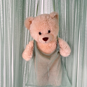 Puede incluir: Un oso de peluche marr&oacute;n claro con ojos marrones y una nariz marr&oacute;n asoma de una bolsa de malla verde claro. La bolsa est&aacute; colgada de un gancho.