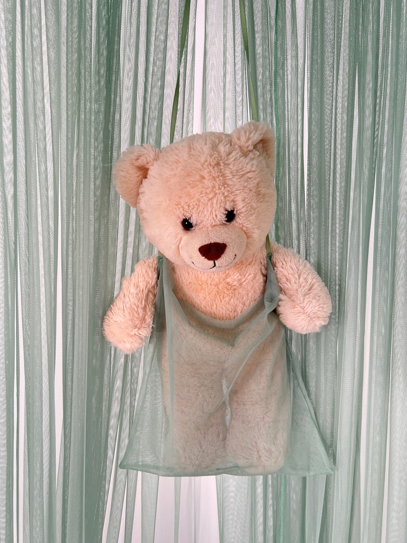 Puede incluir: Un oso de peluche marr&oacute;n claro con ojos marrones y una nariz marr&oacute;n asoma de una bolsa de malla verde claro. La bolsa est&aacute; colgada de un gancho.