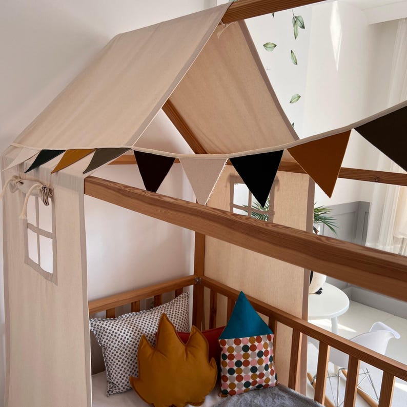 May include: A wooden house-shaped bed frame with a beige canvas roof and side panels. A colorful triangular banner hangs across the front. The bed is decorated with pillows in various shapes and patterns, including a leaf and a house.