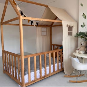 May include: A wooden house-shaped bed frame with a natural finish. The bed has a slatted side rail and a fabric roof and curtains. Decorative pennants hang from the roof. A white mattress and gray blanket are visible.