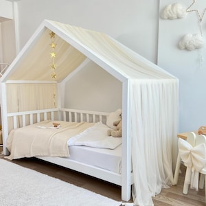 May include: A white wooden house-shaped bed frame featuring a peaked roof draped with sheer cream-coloured fabric. A gold star garland hangs from the top beam. The bed is dressed with a cream-coloured textured blanket and a white pillow.