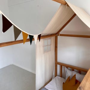 May include: A wooden house-shaped bed frame with a cream-colored canopy and a small window. A garland of colorful pennants hangs above the bed. The bed is decorated with pillows and a white sheet.