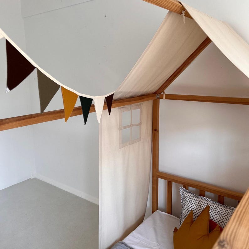 May include: A wooden house-shaped bed frame with a cream-colored canopy and a small window. A garland of colorful pennants hangs above the bed. The bed is decorated with pillows and a white sheet.