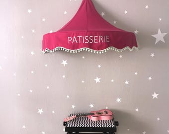 Könnte beinhalten: Ein pinkfarbenes Baldachin mit dem Wort "PATISSERIE" in weißen Buchstaben, verziert mit weißen Pompons, hängt an einer Wand, die mit weißen Sternaufklebern verziert ist. Eine rosa Ukulele liegt auf einer schwarz-weiß gemusterten Bank.
