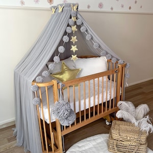 Può includere: Una culla in legno con baldacchino grigio e dettagli decorativi. La culla è adornata con un cuscino a forma di stella dorata, ghirlande di stelle e pompon e una decorazione floreale grigia. Un cestino di vimini e piume sono in primo piano.