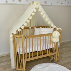 Può includere: Una culla in legno chiaro con materasso e cuscini bianchi. La culla ha un baldacchino in tessuto trasparente ed è decorata con pompon. La culla è su ruote e ha un tappeto rotondo bianco sotto.