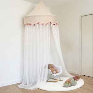 May include: A cream-colored canopy with a scalloped edge adorned with colorful tassels. The canopy hangs from the ceiling with sheer white curtains. A white rocking chair with a stuffed bunny sits on a white rug, with other stuffed toys.