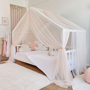May include: A white wooden house-shaped bed frame draped with sheer, pale pink tulle fabric. The bed features a white mattress, a light pink textured blanket, and decorative pillows shaped like a star and clouds in shades of pink and white.
