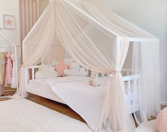 Puede incluir: Una estructura de cama blanca con forma de casa cubierta con tela de tul transparente de color rosa pálido. La cama incluye un colchón blanco, una almohada en forma de estrella rosa claro, dos almohadas de nube y un peluche.