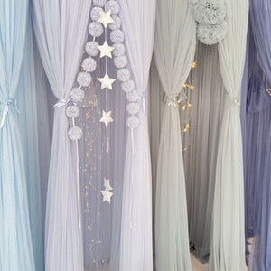 May include: A set of five sheer curtains in various shades of blue, decorated with white pom-poms and star-shaped lights. The curtains are hung from a ceiling and create a whimsical, dreamy atmosphere.