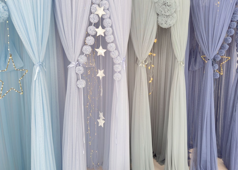 May include: A set of five sheer curtains in various shades of blue, decorated with white pom-poms and star-shaped lights. The curtains are hung from a ceiling and create a whimsical, dreamy atmosphere.