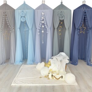May include: Five different color tulle canopy tents with pom poms and star string lights hanging from the top. The tents are set up in a room with a white rug and pillows in front of them.