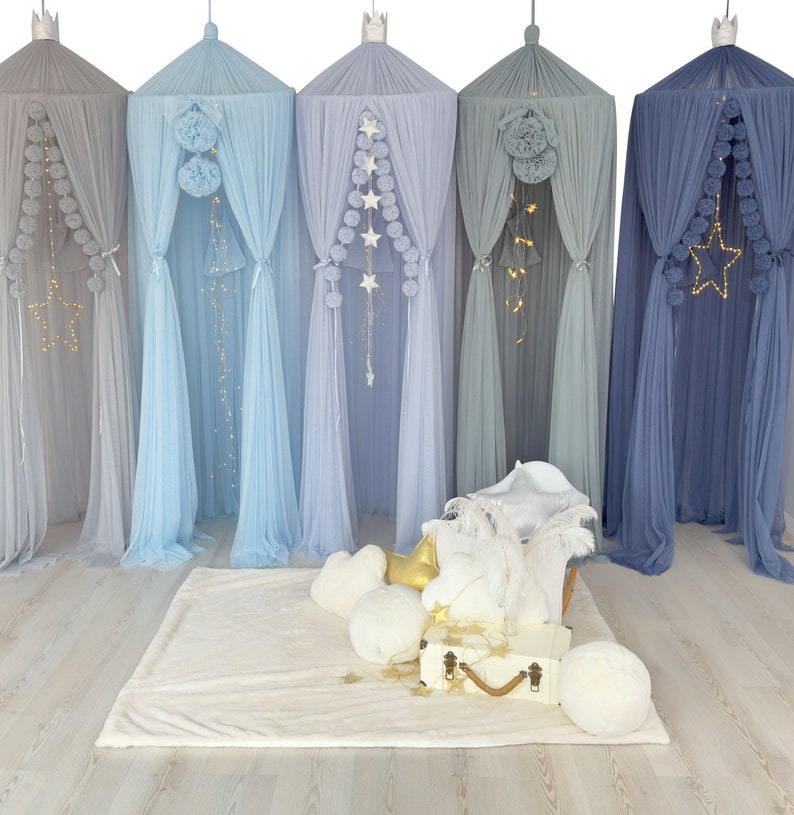 May include: Five different color tulle canopy tents with pom poms and star string lights hanging from the top. The tents are set up in a room with a white rug and pillows in front of them.