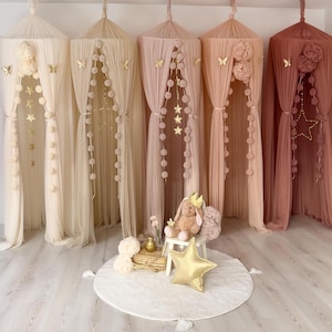 May include: A display of five sheer canopy beds in varying shades of cream, pink, and mauve. Each canopy is adorned with decorative elements like butterflies, stars, and pom-poms. A plush bunny with a crown sits on a small white stool.