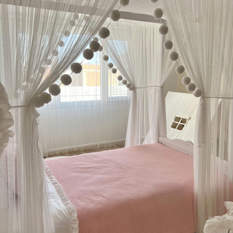 May include: A white canopy bed with a pink blanket and white pom poms strung across the top.