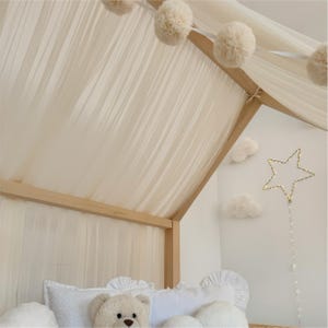 May include: A wooden house-shaped bed frame draped with sheer cream fabric and a string of cream-colored pom-poms. A light-up star decoration and two white cloud-shaped wall accents are visible. A plush teddy bear sits on white pillows.