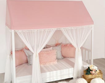 Op de afbeelding: Een wit houten bedframe in de vorm van een huis met een roze stoffen dak en witte kwastjesrand. Het bed is voorzien van doorschijnende witte gordijnen aan de stijlen, roze kussens met ruches en een wit matras op een lattenbodem.