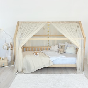 May include: A wooden house-shaped bed frame featuring a light beige tulle canopy draped over the top. The bed is dressed with a white sheet, a beige blanket, two cloud-shaped pillows, and a small teddy bear. A vertical string of gold stars hangs from the center.