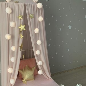 Puede incluir: Una cama con dosel rosa con una guirnalda de estrellas doradas y luces de cadena. La cama tiene una almohada rosa y una almohada dorada en forma de corona. La pared detr&aacute;s de la cama es gris con calcoman&iacute;as de estrellas blancas.